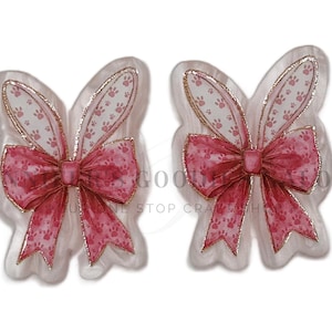 May include: Two bunny ear-shaped embellishments with a pink bow and paw print details. The ears have a gold outline and a sheer, patterned interior. The bows are a solid pink color. These are likely craft supplies or decorative elements.