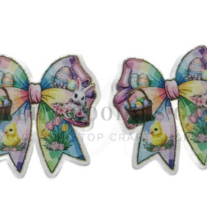 May include: Two Easter-themed bows with a colorful design. Each bow features a basket of eggs, a bunny, and a chick. The bows have a rainbow color scheme with a plaid pattern and are outlined in silver.