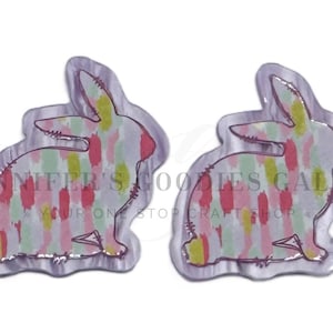 May include: Two bunny-shaped craft embellishments with a watercolor design. The bunnies have a white outline and are filled with vertical strokes of pink, green, and yellow. The text "JENNIFER'S GOODIES GALORE" is visible.