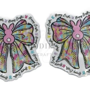 May include: Two Easter-themed bow-shaped embellishments. Each bow features a pink bunny silhouette and a colorful watercolor design. The words "Happy Easter" are written around the edges.