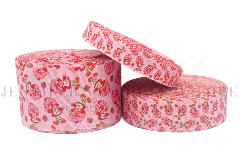 May include: Three rolls of pink ribbon with a repeating pattern of a cartoon strawberry girl and strawberries. The ribbon is a light pink color with a textured surface. The rolls are of varying sizes.