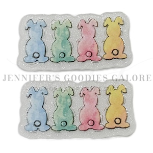 May include: Two sets of Easter-themed embellishments, each featuring eight colorful bunny silhouettes. The bunnies are in pastel shades of blue, green, pink, and yellow, outlined in black, and set against a glittery white background. The text "JENNIFER'S GOODIES GALORE" is visible.