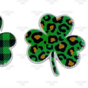 May include: Two shamrock-shaped pins. One has a green and black plaid pattern, the other a green background with a leopard print design in brown and black. A US dollar coin is next to the shamrocks.