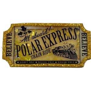 May include: A rectangular ticket-shaped ornament with a gold glitter border. The center features the text All aboard! POLAR EXPRESS TRAIN RIDE, a steam locomotive illustration, holly leaves, and the word BELIEVE on both sides. The bottom reads ADMIT ONE ROUND TRIP NORTH POLE.