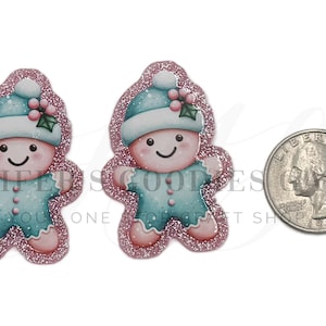 May include: Two gingerbread man-shaped Christmas ornaments with a pink glitter outline. Each ornament has a blue hat with a white pom-pom and holly berries. The gingerbread men are wearing blue outfits and have smiling faces. A quarter is next to the ornaments.