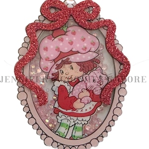 May include: A pink and red glittery framed shaker charm featuring a cartoon character in a strawberry hat holding a pink cat. The charm has a red glitter bow at the top and is filled with small pink glitter pieces.