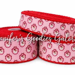 May include: Three spools of light pink grosgrain ribbon featuring a repeating pattern of Santa Claus faces inside red oval frames. The edges of the ribbon are finished with a textured red trim. The text Jennifer's Goodies Galore is overlaid.