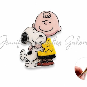 May include: A flat, decorative acrylic charm featuring a cartoon illustration of a bald boy in a yellow shirt with a black zigzag pattern hugging a white dog with black ears and a black spot. The charm has a glittery base and a side profile view.
