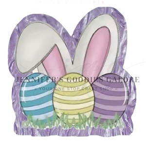 May include: Easter-themed decoration featuring bunny ears and three decorated eggs. The eggs are blue, yellow, and purple, with horizontal stripes. The background is a textured purple, and the image includes the text "JENNIFER'S GOODIES GALORE".