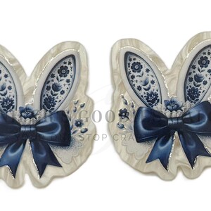 May include: Two bunny-shaped ornaments with a cream-colored base and blue floral designs. Each ornament features a navy blue bow and detailed bunny ears. The ears have a blue and white floral pattern. The overall design is elegant and decorative.