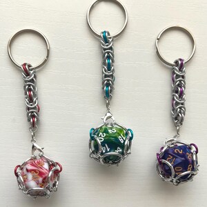 May include: Three keychains with dice inside metal cages. One keychain has a red and white marbled die, another has a green and blue die, and the third has a purple and gold die. Each keychain has a silver ring.