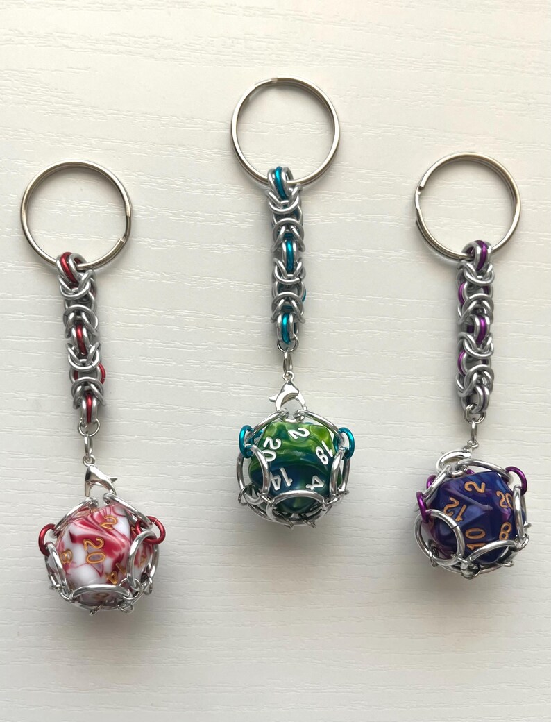 May include: Three keychains with dice inside metal cages. One keychain has a red and white marbled die, another has a green and blue die, and the third has a purple and gold die. Each keychain has a silver ring.