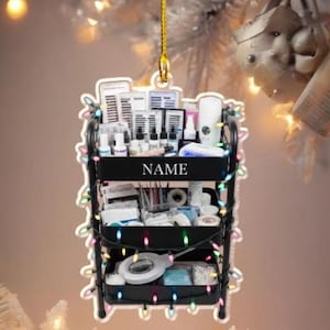 May include: A black miniature rolling cart ornament, decorated with colorful lights. The cart is filled with beauty supplies, including bottles, tools, and lash extensions. The word "NAME" is printed on the cart.