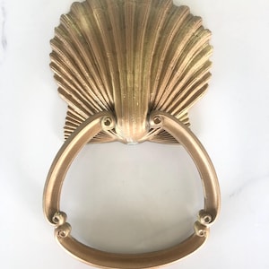 May include: A gold-colored, seashell-shaped door knocker. The knocker features a detailed, fan-like design with a curved handle. The metal has a slightly aged appearance, adding to its vintage charm. The knocker is mounted on a white surface.