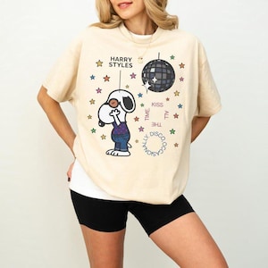 Könnte beinhalten: Beiges kurzärmeliges T-Shirt mit einem Snoopy-ähnlichen Charakter, einer Discokugel und dem Text "Harry Styles", "Kiss All The Time" und "Occasionally Disco". Das Shirt hat einen Rundhalsausschnitt und wird mit schwarzen Shorts kombiniert.