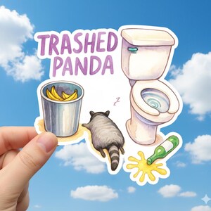 May include: A sticker with the text "Trashed Panda" above a cartoon illustration of a raccoon, a toilet, a trash can with bananas, and a spilled bottle. The sticker is held against a blue sky background.