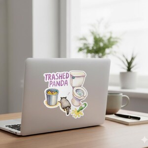 May include: A sticker featuring a cartoon raccoon, a toilet, a bucket of bananas, and a spilled bottle. The text "Trashed Panda" is above the illustrations. The sticker is on a laptop.