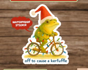 May include: A waterproof sticker featuring a cartoon frog riding a bicycle decorated with Christmas lights. The frog wears a Santa hat and the text "off to cause a kerfuffle" is below. The sticker is on a wooden background.