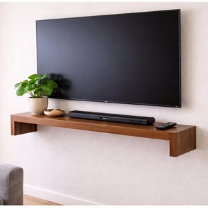 May include: A modern living room setup featuring a large, black flat-screen TV mounted on a white wall. Below the TV is a floating wooden shelf with a soundbar, remote, and decorative items. A potted plant adds a touch of greenery.