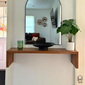 May include: A wooden floating shelf with a black-framed arched mirror above it. A green glass, a black bowl, and a potted plant sit on the shelf. The mirror reflects a living room with a brown sofa and decorative wall shelves.