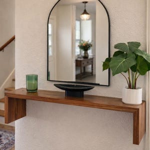 May include: A modern entryway with a wooden console table, a black arched mirror, and a potted plant. A green candle and a black decorative bowl sit on the table. A wooden door and staircase are visible in the background.
