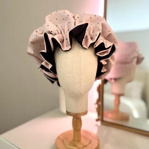 May include: A pink shower cap with black polka dots and a black ruffled trim. The cap is displayed on a mannequin head on a wooden stand. A mirror reflects the cap and stand.