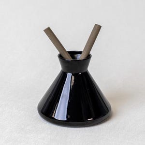 May include: A glossy black ceramic diffuser vessel with a wide, conical base and a narrow neck. Two straight, matte brown wooden reeds are inserted into the opening, protruding upwards in a V-shape. The object has a smooth, reflective surface.