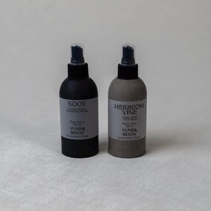 May include: Two cylindrical room spray bottles with black pump nozzles. The left bottle is matte black with a label reading SOOT, while the right bottle is a textured grey concrete finish with a label reading HEIRLOOM VINE. Both feature VUNEA BETON branding.