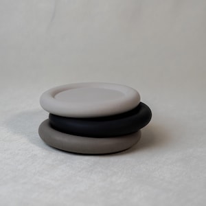 May include: A stack of three round coasters in neutral colors. The top coaster is light gray, the middle one is black, and the bottom one is a darker gray. The coasters are stacked on top of each other.