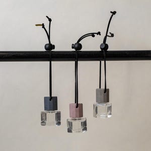May include: Three small, square glass bottles hang from a black horizontal rod by black cords with adjustable black beads. Each bottle features a different colored cap: one dark gray, one dusty pink, and one light gray, all containing clear liquid.