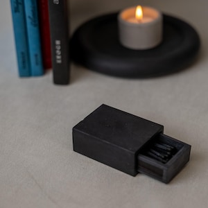 May include: A rectangular matte black matchbox with a sliding tray partially open, revealing several black-tipped matches inside. The matchbox is positioned on a neutral surface, with a lit candle and books visible in the background.