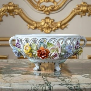 以下が含まれることがあります： 白磁のセンターピースボウル。透かし彫りのデザインと花のアクセントが特徴です。ボウルには、黄色、オレンジ、ピンク、紫の色合いのカラフルな立体的な花が描かれています。3つの小さな足の上に置かれています。