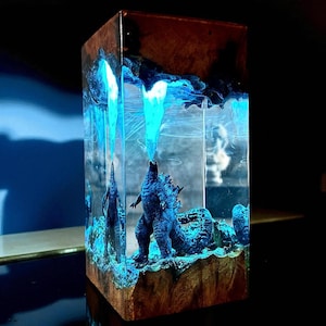 Puede incluir: Escultura rectangular de resina con una figura azul de Godzilla emitiendo un rayo azul. La escultura está encapsulada en resina transparente con detalles azules, sobre una base de madera. El efecto general es una exhibición artística y brillante.