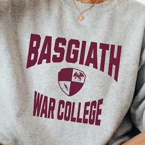 Puede incluir: Una sudadera gris jaspeado con el texto burdeos "BASGIATH WAR COLLEGE" y un escudo. La imagen muestra a una persona sosteniendo una taza negra. El diseño incluye un dragón y un escudo.