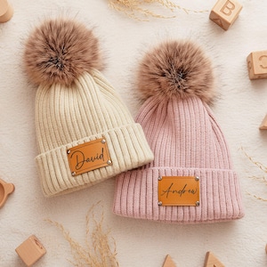 Puede incluir: Dos gorros de punto acanalado, uno crema y otro rosa, cada uno con un pompón marrón. El gorro crema tiene una etiqueta de cuero con "David", y el rosa "Andrew". Juguetes de madera, incluyendo cubos, están alrededor.
