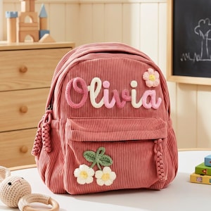 May include: A dusky pink corduroy backpack with the name "Olivia" in a textured font. The backpack has a front pocket and is decorated with floral and cherry appliqués. A small, crocheted bunny toy and wooden ring are on the table.