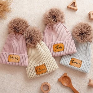 May include: Four ribbed knit beanies in pink, cream, and light blue, each with a faux fur pom-pom. The beanies have a leatherette patch with a name on each: Mary, Clara, Iris, and Sophia. Wooden blocks and toys surround the hats.
