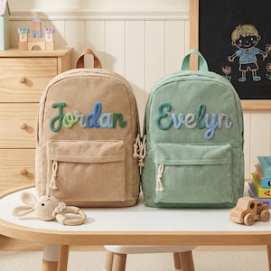 May include: Two corduroy backpacks, one tan and one sage green, sit on a white table. The tan backpack has the name "Jordan" embroidered in green and blue. The green backpack has "Evelyn" embroidered in blue and green.