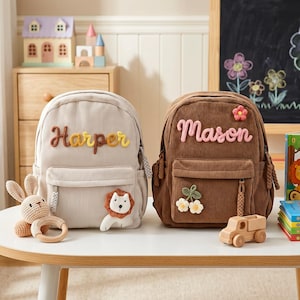 Puede incluir: Dos mochilas de pana, una beige con "Harper" en letras amarillas y un parche de león, la otra marrón con "Mason" en letras rosas y detalles florales. Un camión de juguete de madera y un conejito de peluche están cerca.