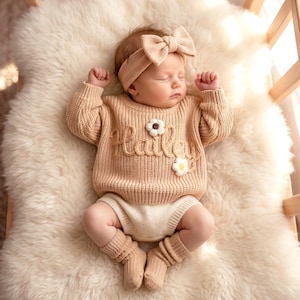 Könnte beinhalten: Ein schlafendes Baby in einem beigefarbenen Strickpullover mit dem Namen "Hailey" und zwei Blumenapplikationen. Das Baby trägt außerdem ein passendes Stirnband, Shorts und Beinwärmer. Das Baby liegt auf einem flauschigen, cremefarbenen Teppich.