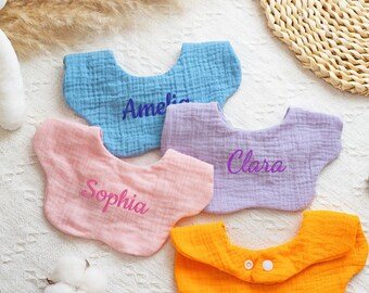May include: Four personalized baby bibs in pastel colors: light blue, pink, lavender, and orange. Each bib has a name embroidered in a matching color: Amelia, Sophia, Clara. The orange bib has two white buttons.