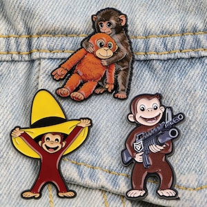 Peut inclure: Trois épingles en émail représentant des singes de dessins animés. L'une montre deux singes s'embrassant, une autre représente un singe portant un sombrero, et la dernière montre un singe tenant un fusil. Les épingles sont posées sur un fond en denim.
