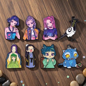 Könnte beinhalten: Eine Sammlung von Emaille-Anstecknadeln mit Anime-Charakteren und -Kreaturen. Die Anstecknadeln zeigen leuchtende Farben und detaillierte Designs, darunter ein Vogel, eine Katze und mehrere weibliche Figuren mit einzigartigen Frisuren und Outfits. Die Anstecknadeln sind auf einer Holzoberfläche angeordnet.