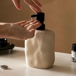 Op de afbeelding: Een beige, getextureerde zeepdispenser met een zwarte pomp. De dispenser is rechthoekig met afgeronde randen. Een hand doseert vloeibare zeep uit de pomp. De dispenser staat op een wit oppervlak.
