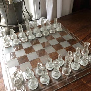 Puede incluir: Un juego de ajedrez de cristal transparente con piezas esmeriladas sobre un tablero de cristal con un patrón de cuadros de madera. Las piezas de ajedrez incluyen peones, torres, caballos, alfiles, una reina y un rey. El juego se exhibe sobre una mesa de madera.