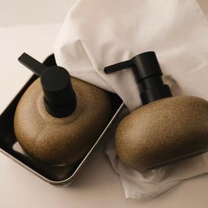 May include: Two brown speckled ceramic soap dispensers with black pumps. One dispenser is in a metal tray, and the other rests on a white cloth. The dispensers have a rounded shape, and the pumps are positioned for easy use.