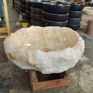 May include: A natural stone sink with a rough, textured exterior and a smooth interior. The sink is a light beige color with streaks of darker hues, and it sits on a wooden support. Other stone sinks are visible in the background.