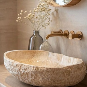 Puede incluir: Un lavabo de piedra natural con un exterior rugoso y texturizado. El lavabo, de forma ovalada, se apoya sobre una superficie de madera. Un grifo de latón y un espejo redondo están montados en la pared. También hay un jarrón con flores secas.