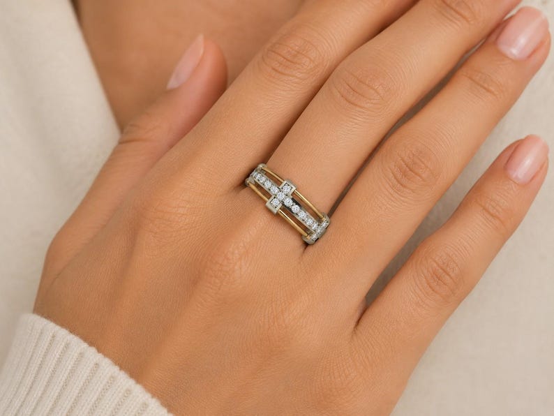 Peut inclure: Une bague bicolore avec une bande dor&eacute;e et un dessus argent&eacute;. La bague pr&eacute;sente un motif en croix avec de petites pierres transparentes. La bague est sur une main &agrave; la peau claire.