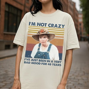 May include: Cream-colored t-shirt with a graphic featuring a woman in a straw hat and overalls. The text "I'M NOT CRAZY" is above the image, and "I'VE JUST BEEN IN A VERY BAD MOOD FOR 40 YEARS" is below. The design has a retro, vintage style.